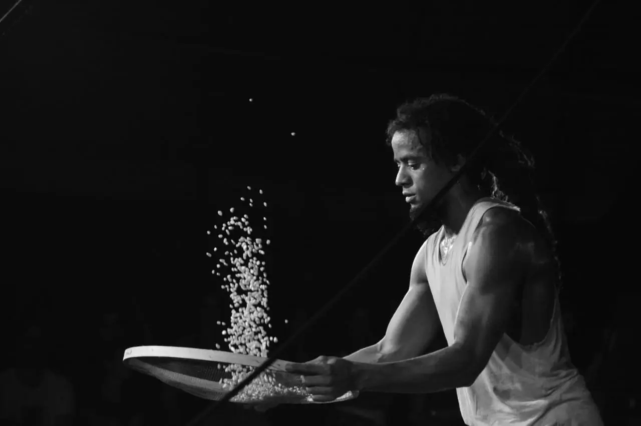 Artista lança grãos ao ar com uma peneira, no escuro do palco